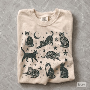 Op de afbeelding: Een ivoorkleurig t-shirt met ronde hals en een zwarte grafische print van zeven katten in verschillende houdingen, waaronder zittend, uitrekkend en lopend, afgewisseld met sterren en een maansikkel. Het binnenlabel vermeldt Comfort Colors.