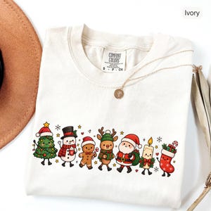 Pode incluir: Uma t-shirt cor de marfim com uma impressão gráfica de personagens de Natal em estilo cartoon, incluindo uma árvore, boneco de neve, boneco de gengibre, rena, Pai Natal, uma vela e uma meia, com etiqueta Comfort Colors tamanho S.