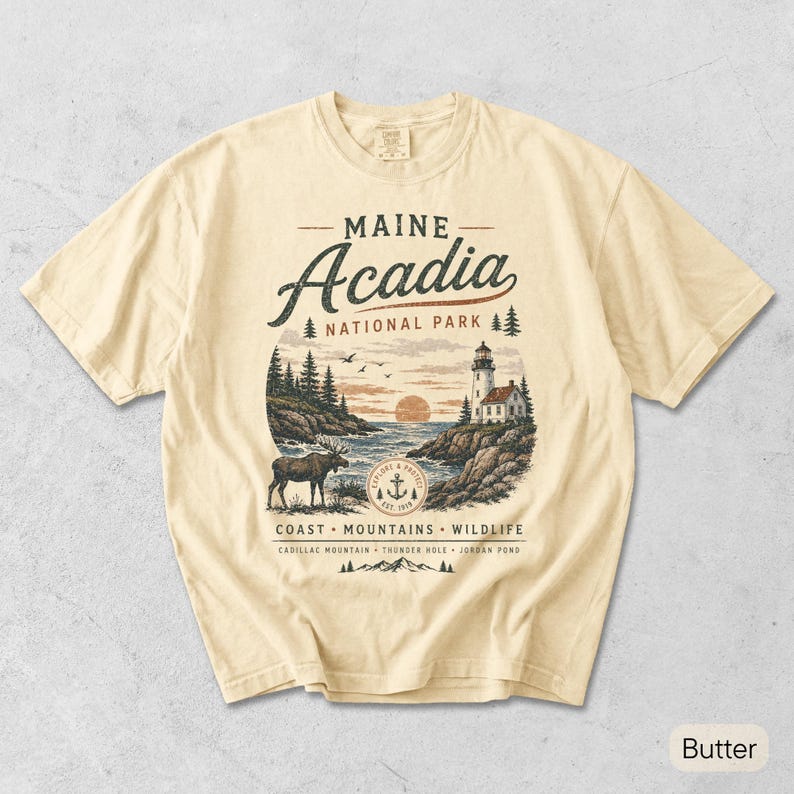 May include: A butter-colored t-shirt featuring a graphic of Maine Acadia National Park. The design includes a lighthouse, a moose, pine trees, and the text "MAINE Acadia NATIONAL PARK COAST MOUNTAINS WILDLIFE CADILLAC MOUNTAIN THUNDER HOLE JORDAN POND".