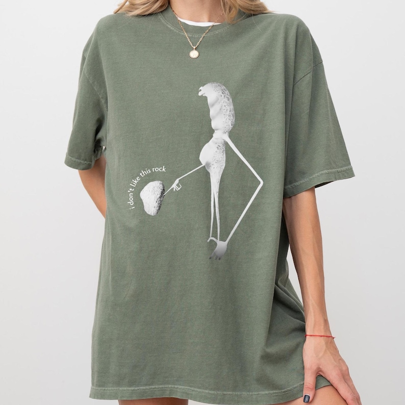 May include: Olive green t-shirt featuring a unique graphic design. The design includes a stylized figure with a rock and the text "I don't like this rock". The shirt has short sleeves and a relaxed fit.