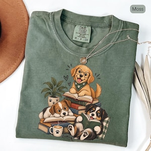 Puede incluir: Una camiseta verde musgo con un gráfico de tres cachorros con gafas, leyendo libros y sentados junto a una planta en maceta y una taza de café. La camiseta tiene una etiqueta de la marca Comfort Colors en el cuello y un collar dorado fino encima.