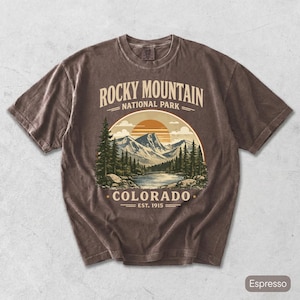 Puede incluir: Camiseta color espresso con un diseño gráfico del Parque Nacional de las Montañas Rocosas en Colorado. El diseño incluye montañas, un lago, árboles y una puesta de sol. El texto en la camiseta dice "Rocky Mountain National Park - Colorado - Est. 1915."