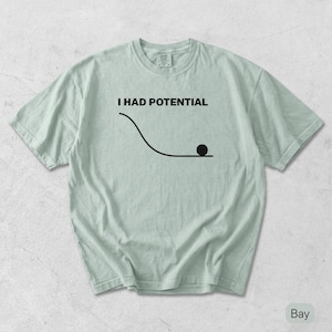 Pode incluir: Uma t-shirt verde s&aacute;lvia claro com uma impress&atilde;o gr&aacute;fica preta. O design inclui o texto "I HAD POTENTIAL" acima de um desenho de linha de uma rampa curva com um pequeno c&iacute;rculo preto a descansar na parte inferior da sec&ccedil;&atilde;o plana.