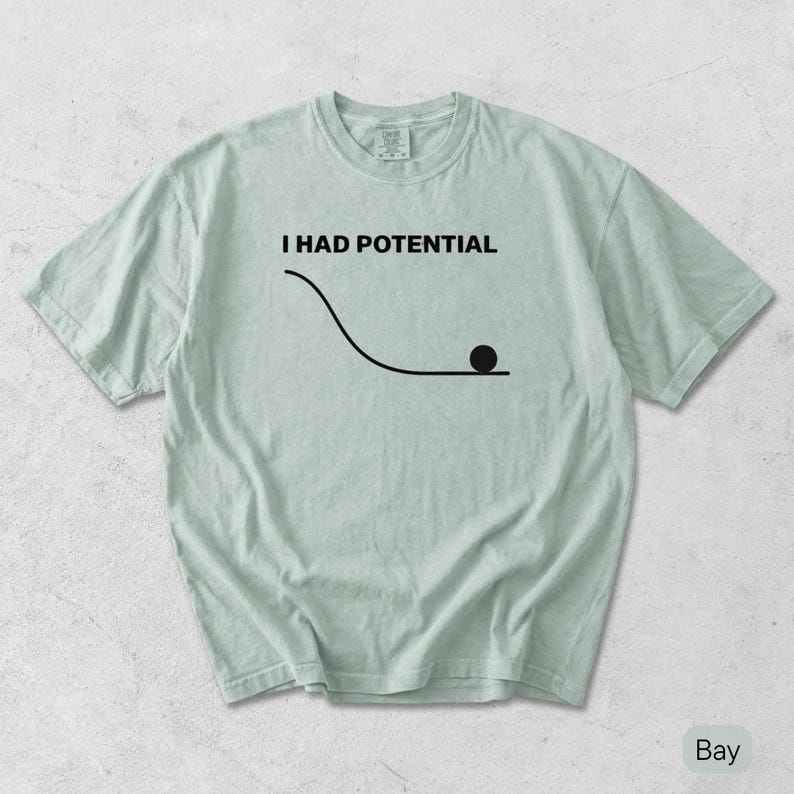 Pode incluir: Uma t-shirt verde s&aacute;lvia claro com uma impress&atilde;o gr&aacute;fica preta. O design inclui o texto "I HAD POTENTIAL" acima de um desenho de linha de uma rampa curva com um pequeno c&iacute;rculo preto a descansar na parte inferior da sec&ccedil;&atilde;o plana.
