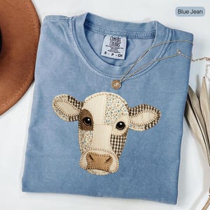 Pode incluir: Camiseta azul jeans com um design de rosto de vaca em patchwork. O rosto da vaca apresenta detalhes em tecido marrom, creme e estampado. Um colar de ouro com um pingente é drapeado sobre a camisa. A camisa tem uma etiqueta Comfort Colors.
