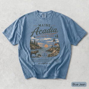May include: A blue jean colored short-sleeve t-shirt featuring a graphic print of Maine Acadia National Park. The design includes a lighthouse, a moose, pine trees, and a sunset over the water with the text Maine Acadia National Park.