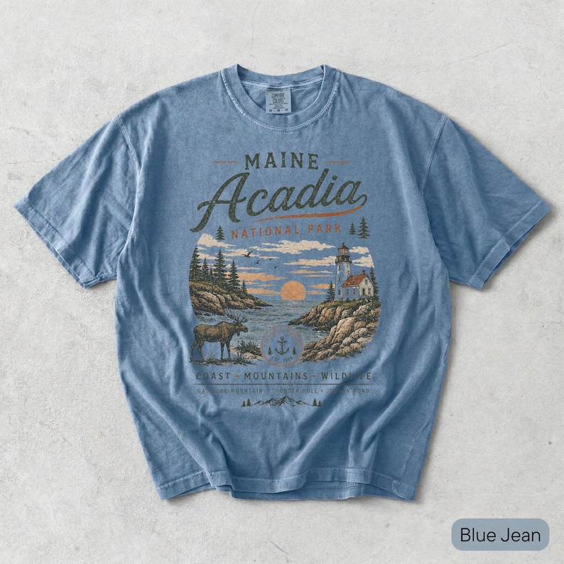 May include: A blue jean colored short-sleeve t-shirt featuring a graphic print of Maine Acadia National Park. The design includes a lighthouse, a moose, pine trees, and a sunset over the water with the text Maine Acadia National Park.