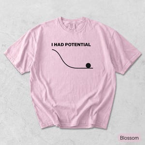 Pode incluir: Uma t-shirt de manga curta em rosa claro com um gr&aacute;fico preto de uma linha curva e um c&iacute;rculo na parte inferior. Acima do gr&aacute;fico, o texto I HAD POTENTIAL est&aacute; escrito em letras mai&uacute;sculas pretas. A t-shirt tem gola redonda.