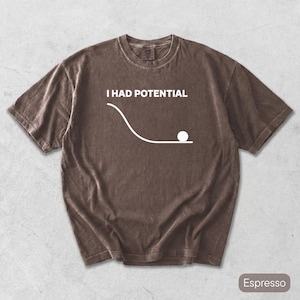 Pode incluir: Uma t-shirt castanha de manga curta com um gr&aacute;fico branco de uma bola na base de uma rampa curva e o texto I HAD POTENTIAL impresso por cima. A camisola tem um decote redondo e um corte descontra&iacute;do numa cor castanha s&oacute;lida.