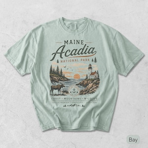 May include: A light sage green t-shirt featuring a graphic of Maine Acadia National Park. The design includes a lighthouse, a moose, pine trees, and the text Maine Acadia National Park, Coast, Mountains, Wildlife, Cadillac Mountain, Thunder Hole, Jordan Pond.