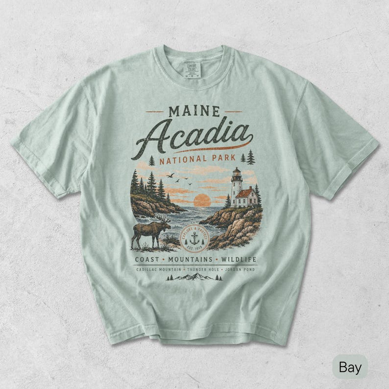 May include: A light sage green t-shirt featuring a graphic of Maine Acadia National Park. The design includes a lighthouse, a moose, pine trees, and the text Maine Acadia National Park, Coast, Mountains, Wildlife, Cadillac Mountain, Thunder Hole, Jordan Pond.
