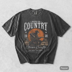 Op de afbeelding: Donkergrijs t-shirt met een vintage afbeelding. De afbeelding toont de tekst "RAISED ON COUNTRY" en een illustratie van een gitaarspelende cowboy. Daaronder staat "Classic Country, REAL MUSIC. REAL STORIES."