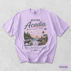 May include: A light purple t-shirt featuring a graphic of Maine Acadia National Park. The design includes a lighthouse, a moose, pine trees, and the text Maine Acadia National Park, Coast, Mountains, Wildlife, Cadillac Mountain, Thunder Hole, Jordan Pond.