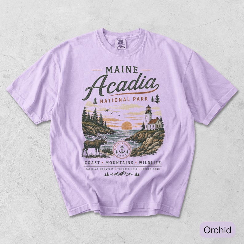 May include: A light purple t-shirt featuring a graphic of Maine Acadia National Park. The design includes a lighthouse, a moose, pine trees, and the text Maine Acadia National Park, Coast, Mountains, Wildlife, Cadillac Mountain, Thunder Hole, Jordan Pond.