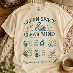 May include: Cream-colored t-shirt with the text "CLEAN SPACE CLEAR MIND" in teal. The shirt features cleaning-themed illustrations and phrases like "Clean today, smile tomorrow" and "Fresh home, fresh vibes."