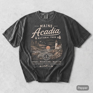 May include: A charcoal gray short-sleeve t-shirt featuring a graphic of Acadia National Park in Maine. The design includes a lighthouse, a moose, a sun, and text reading Maine Acadia National Park, Coast, Mountains, Wildlife, Cadillac Mountain, Thunder Hole, Jordan Pond.