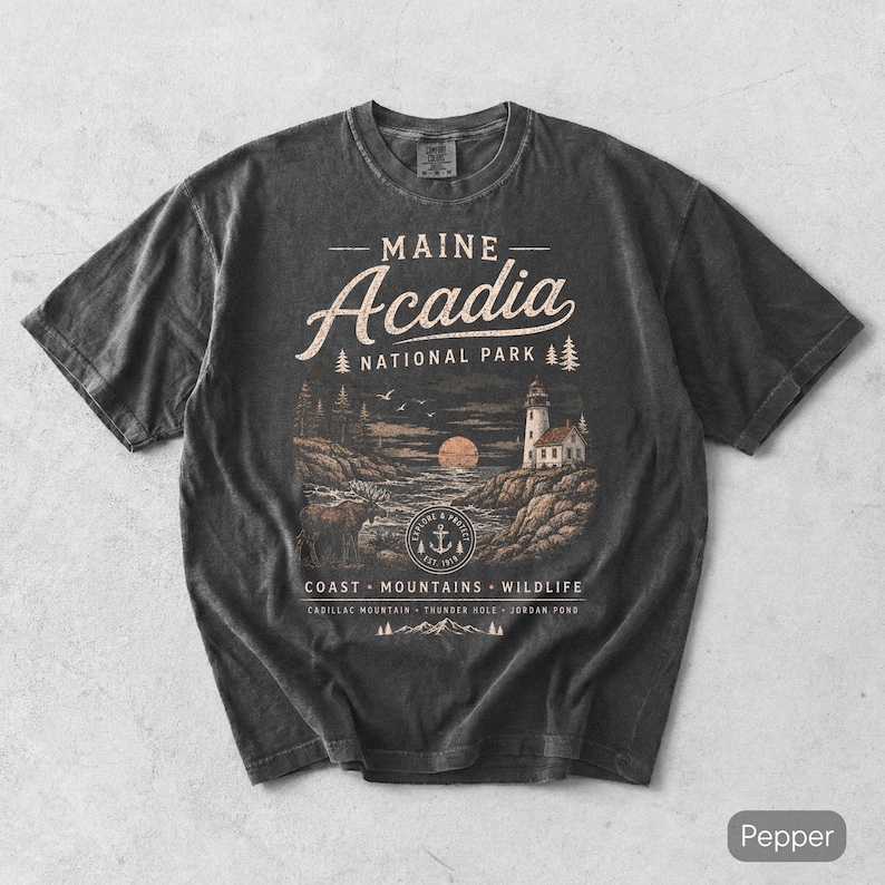 May include: A charcoal gray short-sleeve t-shirt featuring a graphic of Acadia National Park in Maine. The design includes a lighthouse, a moose, a sun, and text reading Maine Acadia National Park, Coast, Mountains, Wildlife, Cadillac Mountain, Thunder Hole, Jordan Pond.