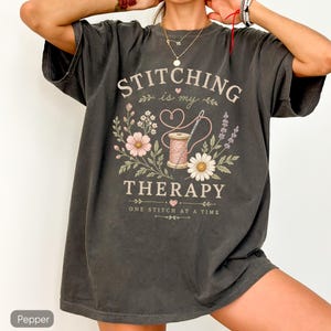 May include: A charcoal gray short-sleeve t-shirt featuring a graphic print with the text "STITCHING is my THERAPY" and "ONE STITCH AT A TIME". The design includes a spool of pink thread, a needle, and various floral illustrations in soft colors.