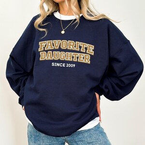 May include: Navy blue sweatshirt with the text "FAVORITE DAUGHTER SINCE 2009" in a gold and brown collegiate style font. The sweatshirt has a white crew neck collar and is a relaxed fit.