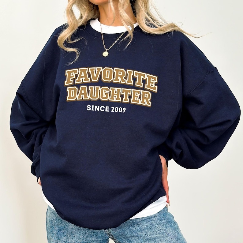 May include: Navy blue sweatshirt with the text "FAVORITE DAUGHTER SINCE 2009" in a gold and brown collegiate style font. The sweatshirt has a white crew neck collar and is a relaxed fit.