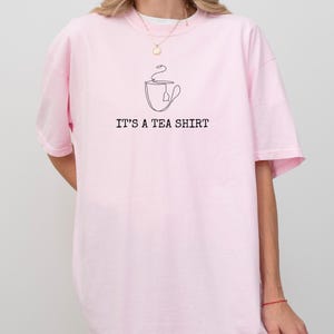 May include: A light pink t-shirt with the text "IT'S A TEA SHIRT" in black. Above the text is a line drawing of a teacup with a tea bag. The shirt is a casual style.