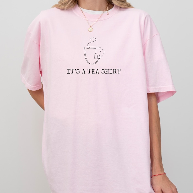 May include: A light pink t-shirt with the text "IT'S A TEA SHIRT" in black. Above the text is a line drawing of a teacup with a tea bag. The shirt is a casual style.