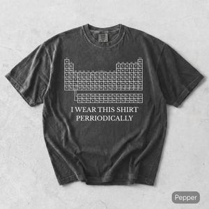Puede incluir: Una camiseta de manga corta en color gris carbón con un gráfico blanco de la tabla periódica de los elementos. Debajo de la tabla, el texto dice I WEAR THIS SHIRT PERRIODICALLY en una fuente serif blanca. La tela tiene un aspecto vintage lavado.