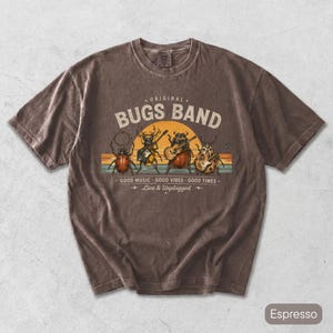 Op de afbeelding: Een espresso-kleurig t-shirt met korte mouwen en een afbeelding van vier kevers die muziekinstrumenten bespelen. De tekst ORIGINAL BUGS BAND, GOOD MUSIC, GOOD VIBES, GOOD TIMES en Live & Unplugged staat op een achtergrond in zonsondergangkleuren.