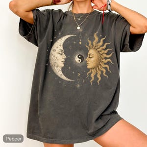 May include: A charcoal gray short-sleeve t-shirt featuring a large graphic print of a crescent moon and a sun face positioned opposite each other. Between the celestial figures is a small yin-yang symbol surrounded by stars and thin line art.