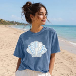 Könnte beinhalten: Ein blaues T-Shirt mit einem Muschelmuster. Die Muschel besteht aus verschiedenen blau-weißen Mustern, darunter Streifen, Vichy-Karo und Blumendrucke. Das T-Shirt hat eine lockere Passform und kurze Ärmel.