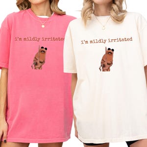 May include: Two t-shirts, one in pink and one in cream, displaying the text "I'm mildly irritated" in brown. Each shirt has a cartoon illustration of a brown creature wearing sunglasses below the text.
