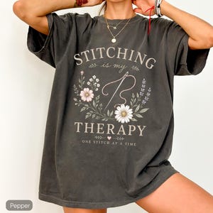 May include: Dark grey t-shirt featuring the text "STITCHING is my THERAPY" within a floral design. The design includes a needle and thread, flowers, and the phrase "ONE STITCH at a TIME".