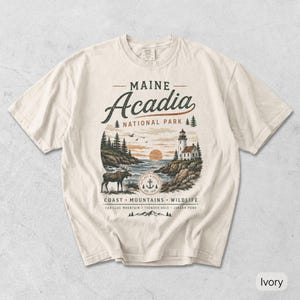 May include: An ivory short-sleeve t-shirt featuring a graphic of Acadia National Park in Maine. The design includes a lighthouse, a moose, a sunset, and text reading Maine Acadia National Park, Coast, Mountains, Wildlife, Cadillac Mountain, Thunder Hole, Jordan Pond.