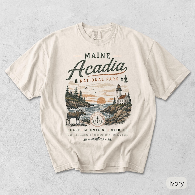 May include: An ivory short-sleeve t-shirt featuring a graphic of Acadia National Park in Maine. The design includes a lighthouse, a moose, a sunset, and text reading Maine Acadia National Park, Coast, Mountains, Wildlife, Cadillac Mountain, Thunder Hole, Jordan Pond.