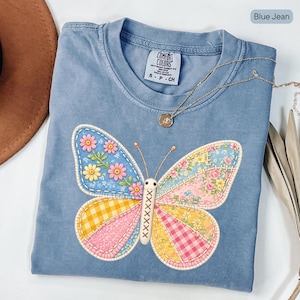 Op de afbeelding: Blauw spijkerbroekkleurig T-shirt met een grote vlinderapplicatie. De vlindervleugels zijn patchwork met bloemen-, ruit- en effen patronen in roze, geel en blauw. Een gouden ketting met een hanger ligt op het shirt.
