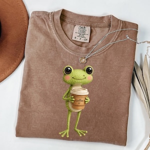 May include: A brown Comfort Colors t-shirt featuring a cartoon frog holding a coffee cup. The frog is green with large eyes and pink cheeks. The coffee cup has the word "coffee" printed on it. A silver necklace is draped across the shirt.