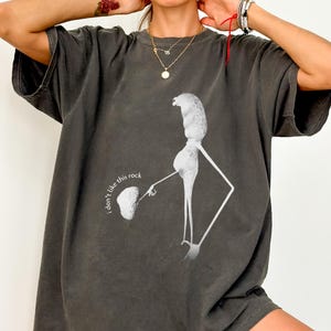 Puede incluir: Camiseta gris oscuro con un gráfico blanco de una figura sosteniendo una roca, con el texto "I don't like this rock." La camiseta tiene mangas cortas y un corte holgado. El diseño está impreso en la parte delantera.