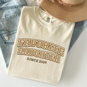 May include: Cream-colored t-shirt with the text "FAVORITE DAUGHTER SINCE 2009" in brown and white lettering. The shirt is folded and placed on top of blue jeans, a necklace, and a tan hat.