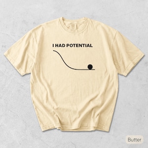 Pode incluir: Uma t-shirt de manga curta na cor manteiga com um gr&aacute;fico preto de uma linha curva e um pequeno c&iacute;rculo na parte inferior, acompanhado pelo texto "I HAD POTENTIAL" impresso em letras mai&uacute;sculas pretas no peito da pe&ccedil;a.