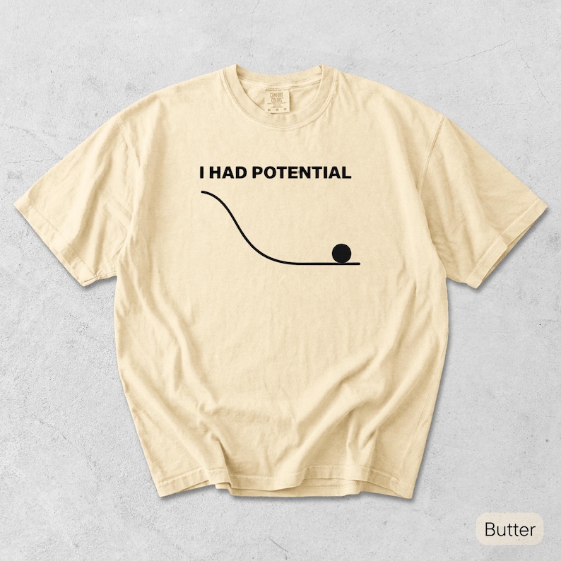 Pode incluir: Uma t-shirt de manga curta na cor manteiga com um gr&aacute;fico preto de uma linha curva e um pequeno c&iacute;rculo na parte inferior, acompanhado pelo texto "I HAD POTENTIAL" impresso em letras mai&uacute;sculas pretas no peito da pe&ccedil;a.
