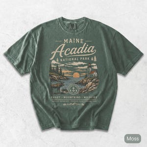 May include: A moss green short-sleeve t-shirt featuring a graphic print of Acadia National Park in Maine. The design includes a lighthouse, a moose, a sunset, and text reading Maine Acadia National Park, Coast, Mountains, Wildlife, Cadillac Mountain, Thunder Hole, Jordan Pond.