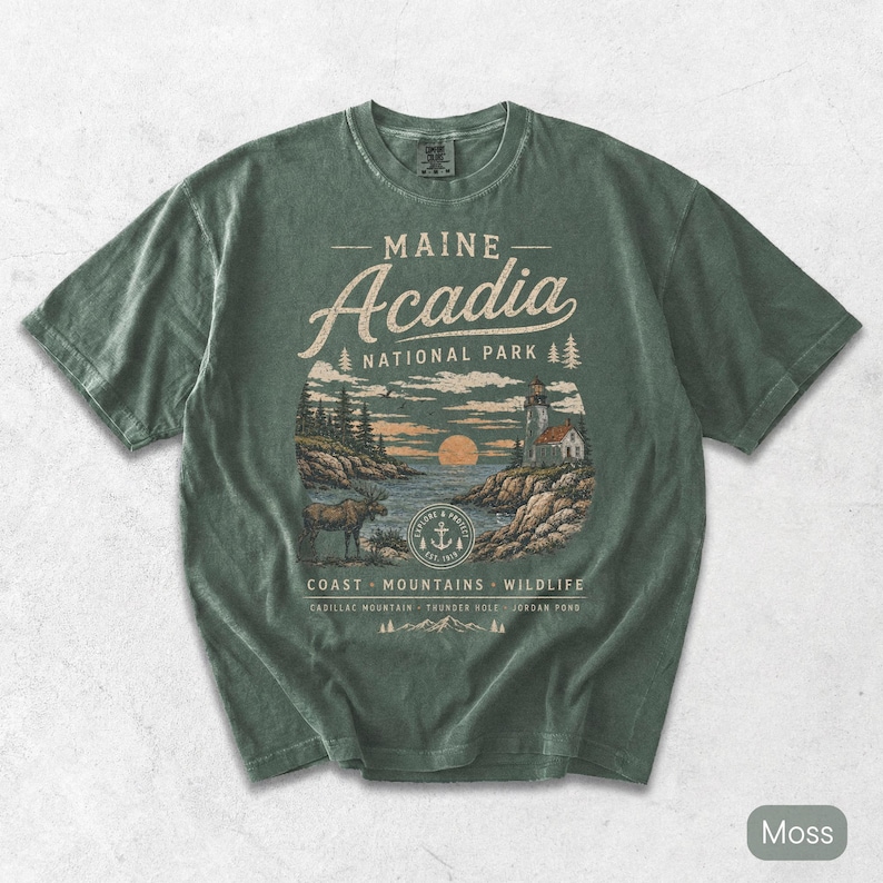 May include: A moss green short-sleeve t-shirt featuring a graphic print of Acadia National Park in Maine. The design includes a lighthouse, a moose, a sunset, and text reading Maine Acadia National Park, Coast, Mountains, Wildlife, Cadillac Mountain, Thunder Hole, Jordan Pond.