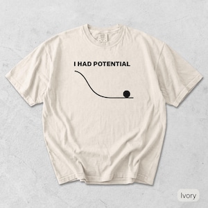 Pode incluir: Uma t-shirt de manga curta na cor marfim com um gr&aacute;fico preto de uma linha curva que leva a uma superf&iacute;cie plana com um c&iacute;rculo preto s&oacute;lido no final. Acima do gr&aacute;fico, o texto I HAD POTENTIAL est&aacute; escrito numa fonte preta, negrito e sem serifa.