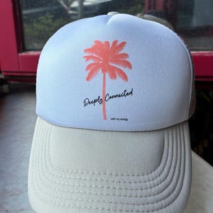 Könnte beinhalten: Zweifarbige Trucker-Mütze mit hellblauem Oberteil und beigem Schirm. Auf der hellblauen Fläche befindet sich eine korallenfarbene Palmen-Grafik und der Text "Deeply Connected with my energy" in schwarzer Schrift.
