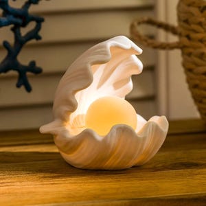 Könnte beinhalten: Muschelförmige Lampe aus cremefarbener Keramik. Im Inneren befindet sich eine leuchtende, kugelförmige Lichtquelle, die ein warmes, einladendes Leuchten erzeugt. Die Lampe steht auf einer Holzoberfläche und erinnert an ein Küsten- oder maritimes Thema.