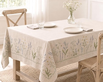 May include: A rectangular beige linen tablecloth featuring a printed botanical pattern of wildflowers in shades of blue, green, and yellow along the edges. The tablecloth is draped over a wooden table with two chairs and a glass vase of flowers.