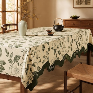 May include: A rectangular tablecloth featuring a cream-colored base with a dark green floral pattern and a scalloped edge. The fabric displays detailed botanical illustrations of flowers and leaves, finished with a solid dark green border trim.