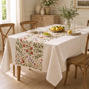 Könnte beinhalten: Eine rechteckige, cremeweiße Tischdecke mit einem mittigen Läuferdesign, das einen farbenfrohen botanischen Druck von Granatäpfeln, Oliven, Lavendel und verschiedenen Wildblumen zeigt. Der Stoff hat eine sichtbare Webstruktur und fällt über einen Esstisch.
