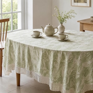 May include: An oval tablecloth with a light green and white botanical pattern and a fringed edge. On the table sits a ceramic teapot, two matching teacups on saucers, and a glass vase containing a bouquet of small white flowers and green stems.