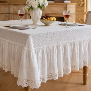 Pode incluir: Uma toalha de mesa quadrada branca com um padrão de diamante texturizado na superfície superior, uma saia larga com babados e um intrincado acabamento em renda ao longo da borda inferior. O tecido é branco sólido com bordados e renda vazada.